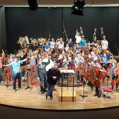 Ein großes Jugend-Sinfonie-Orchester steht in legerer Kleidung auf einer Bühne, alle halten ihre Instrumente hoch in die Luft; vorn steht lächelnd der Orchesterleiter ans Dirigentenpult gelehnt