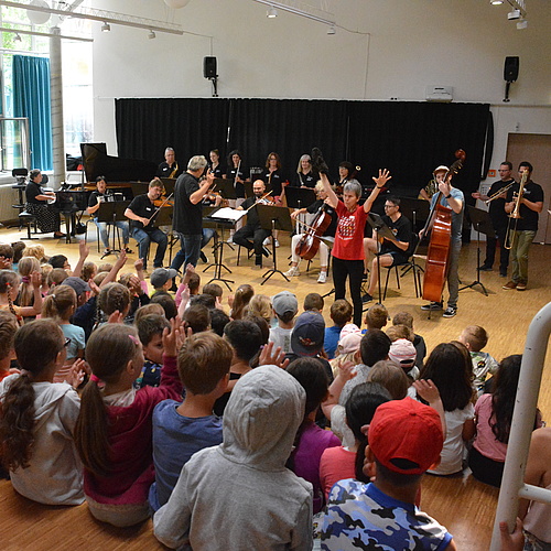 Ein Orchester aus etwa 10 Erwachsenen spielt vor eine größeren Gruppe von Kindern.