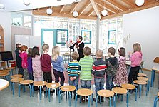 12 Kinder eines Grundschulchors stehen im Halbkreis um die dirigierende und vorsingende Lehrerin